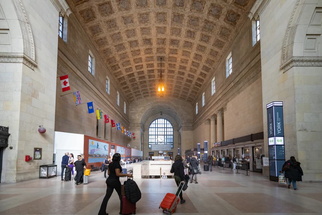 Toronto के Union Station के Great Hall से सूटकेस लेकर गुज़रते यात्री, ऊपर मेहराबदार छत और झंडों की एक क़तार